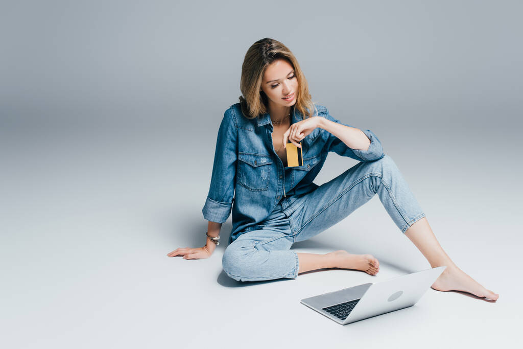 mujer descalza en camisa de mezclilla y jeans con tarjeta de crédito mientras está sentado en gris cerca de la computadora portátil - Foto, Imagen
