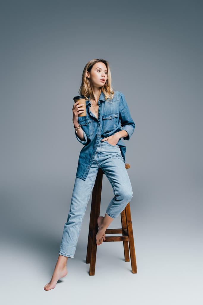 stylish woman in denim clothes sitting on high chair with disposable cup on grey - Photo, Image