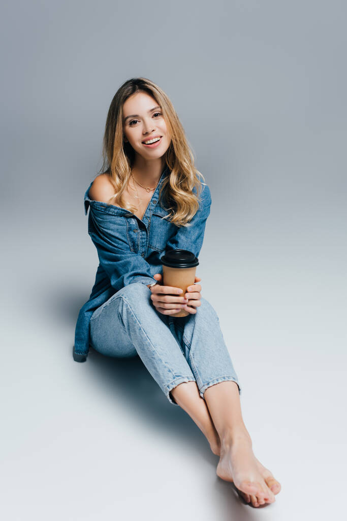 barefoot woman wearing denim clothes, with naked shoulder, sitting with paper cup on grey background - Photo, Image