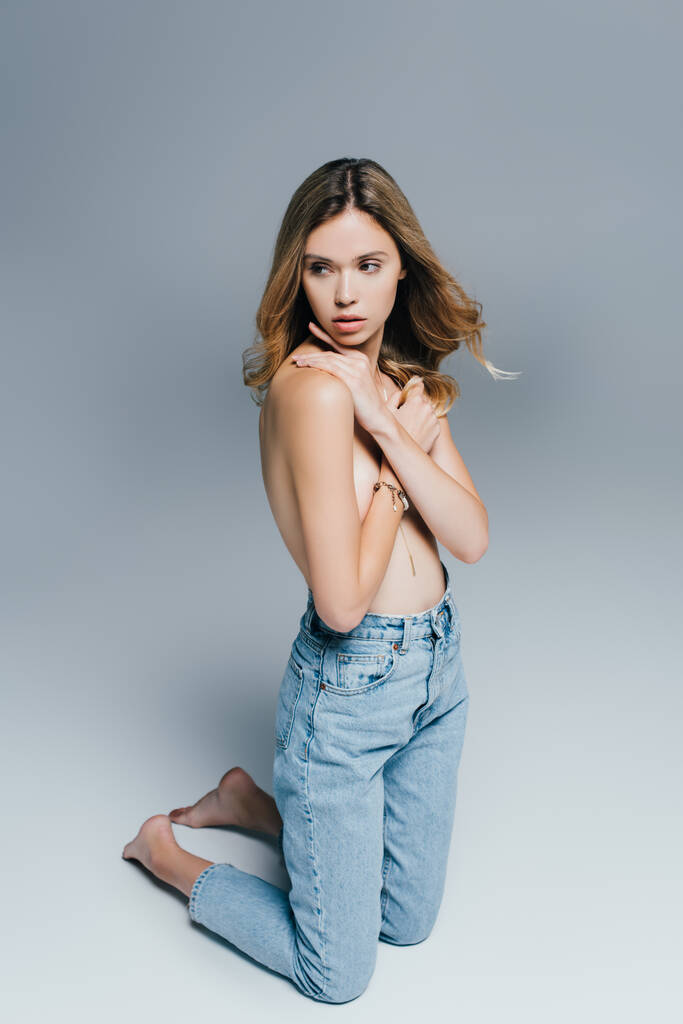 seductive topless woman in jeans hiding breasts with hands while kneeling on grey - Photo, Image