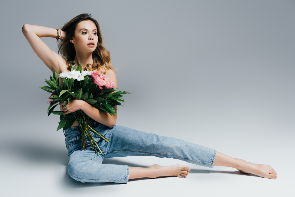 sexy, barefoot woman in jeans, with naked shoulders, touching hair while sitting with flowers on grey - Photo, Image