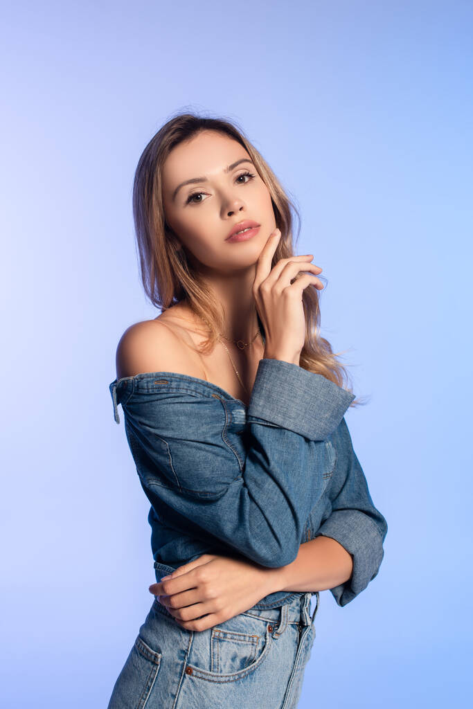 charming young woman in denim shirt, with naked shoulder, touching face isolated on blue - Photo, Image