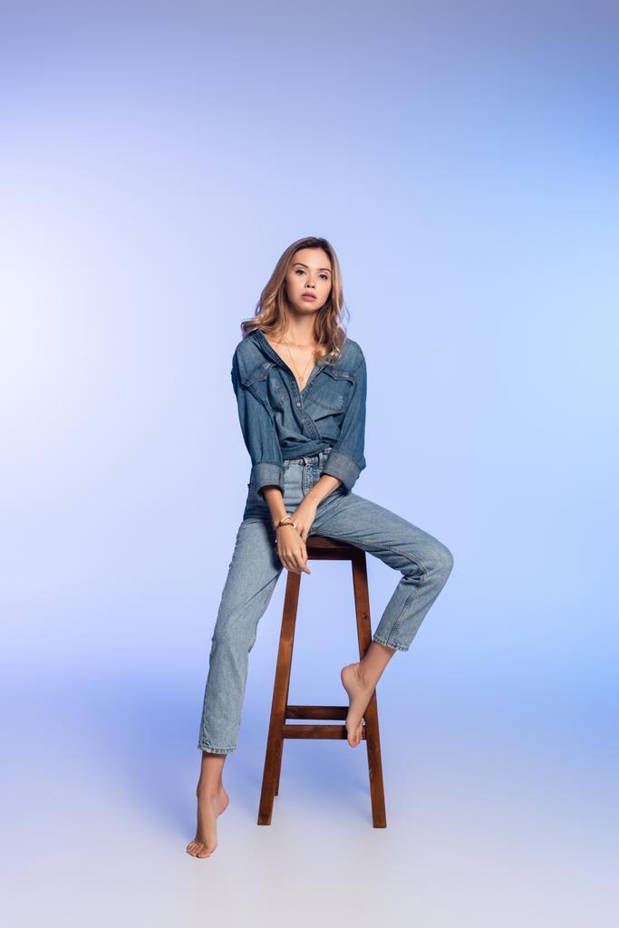 молодая босиком женщина в джинсовой одежде сидит на высоком стуле на голубом - Фото, изображение