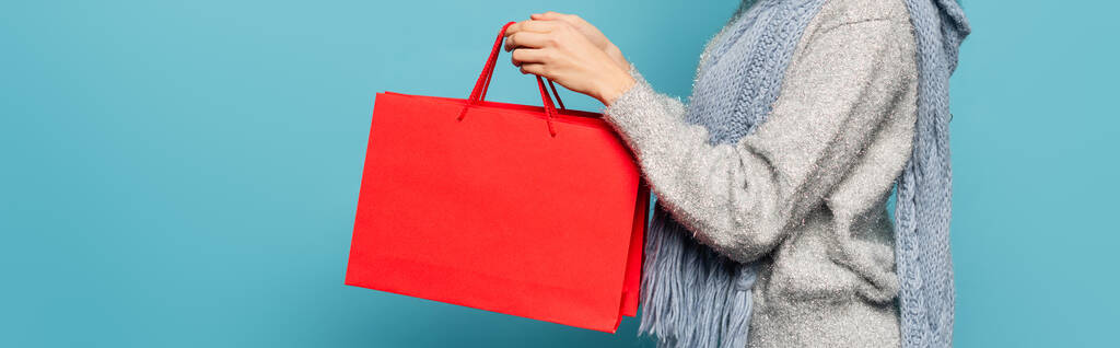 スカーフやセーターを着た女性の作物のビュー、青、バナーに隔離された赤いショッピングバッグを保持 - 写真・画像