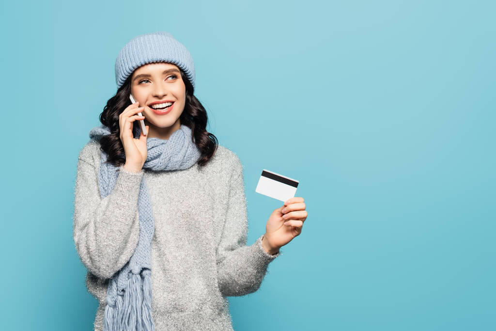 gelukkig brunette vrouw in sjaal en hoed praten op smartphone terwijl het houden van credit card geïsoleerd op blauw - Foto, afbeelding