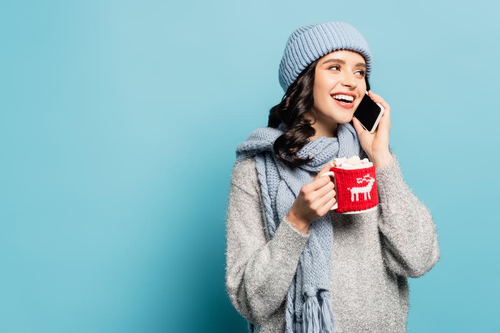 Щаслива жінка в шарфі і капелюсі розмовляє на смартфоні, тримаючи чашку з зефіром і тримачем ізольовані на синьому
 - Фото, зображення