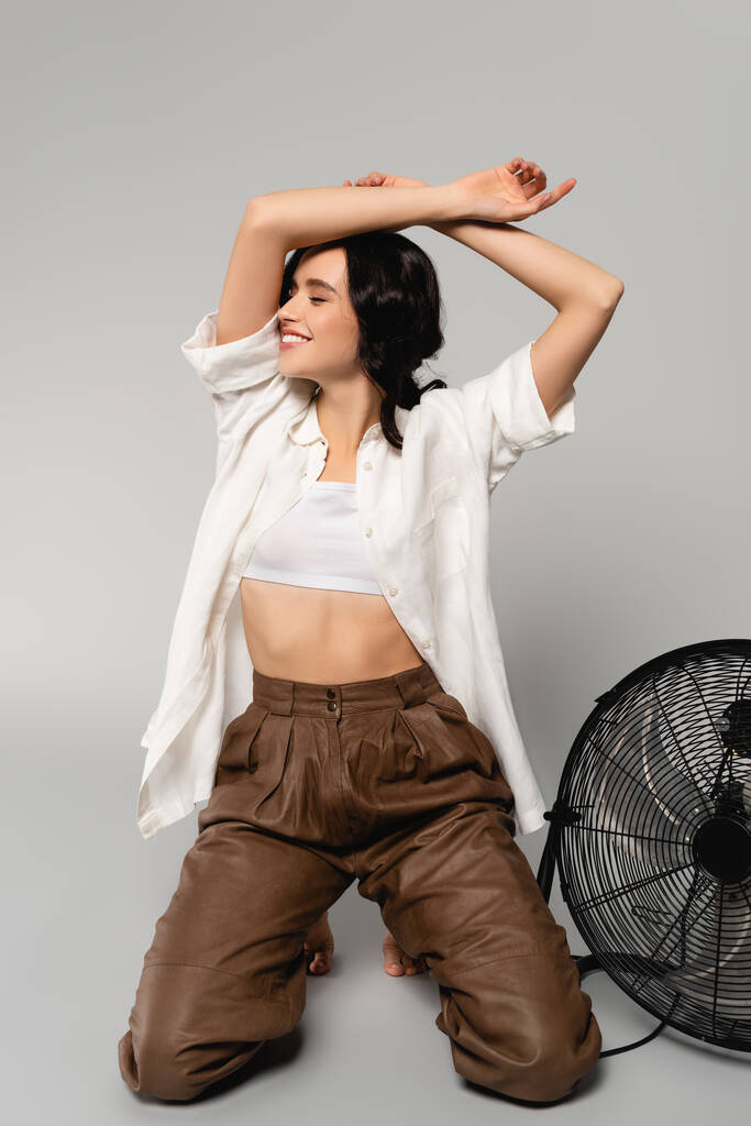 Femme brune souriante avec les mains dans l'air, debout sur les genoux près du ventilateur sur le gris - Photo, image