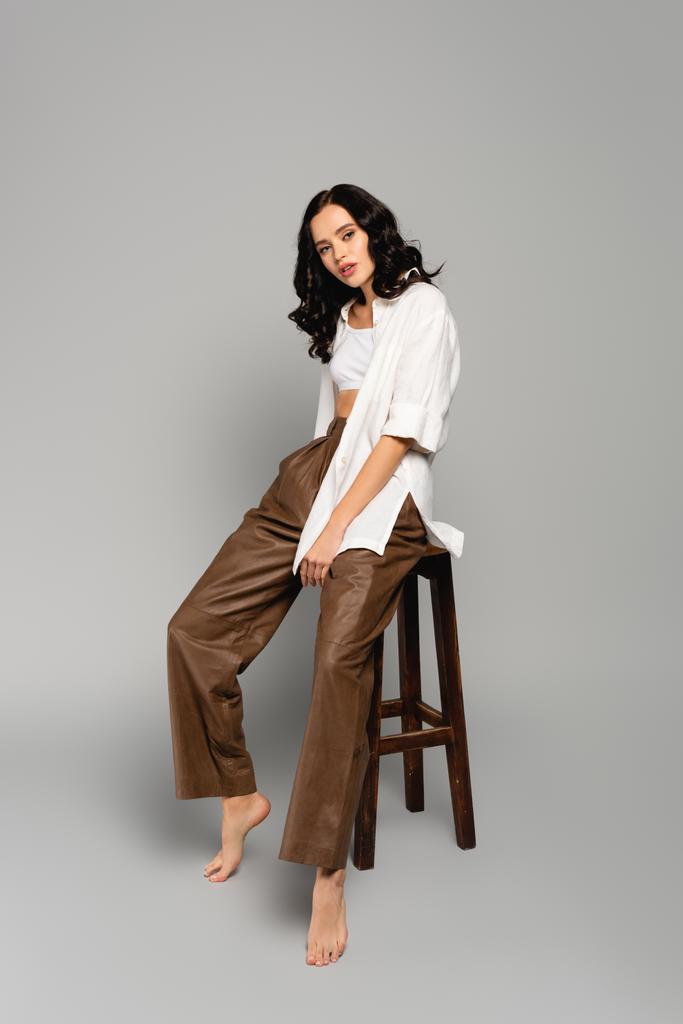 Full length of curly brunette woman in shirt and leather pants posing while sitting on stool on grey background - Photo, Image