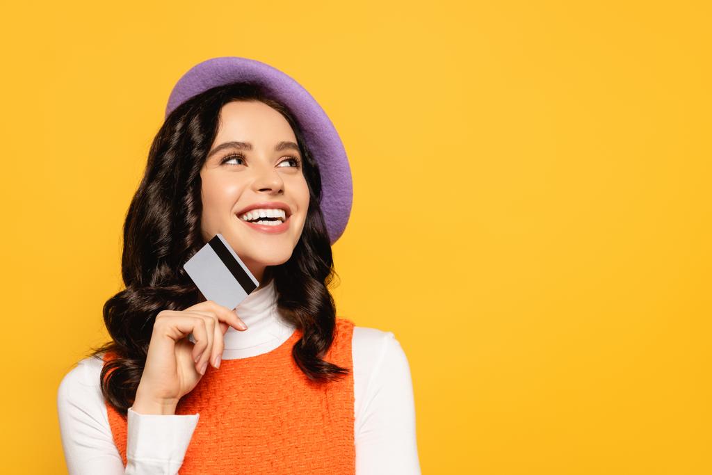 Fröhliche brünette Frau mit Baskenmütze schaut auf, während sie ihre Kreditkarte isoliert auf gelb hält - Foto, Bild