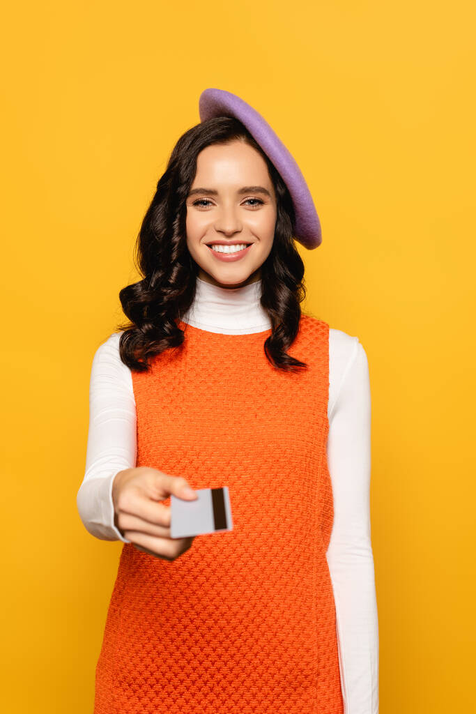 Vooraanzicht van glimlachende brunette vrouw in baret kijken naar camera terwijl het geven van credit card geïsoleerd op geel op wazig voorgrond - Foto, afbeelding