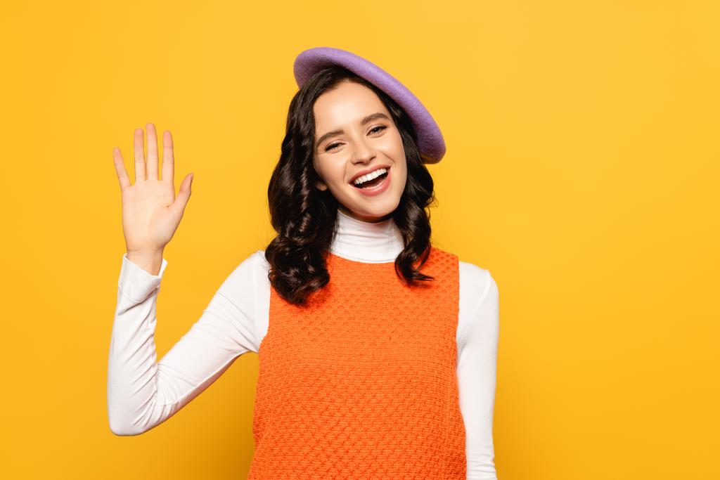 Cheerful brunette woman in beret with waving hand looking at camera isolated on yellow - Photo, Image