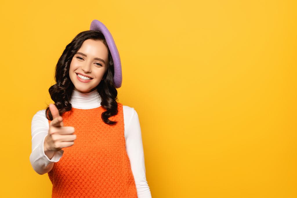 Smiling brunette woman in beret pointing with finger while looking at camera isolated on yellow on blurred foreground - Photo, Image