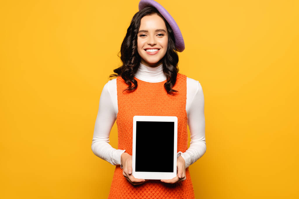 Přední pohled šťastné brunetky žena v baretu při pohledu do kamery, zatímco drží digitální tablet izolované na žluté - Fotografie, Obrázek