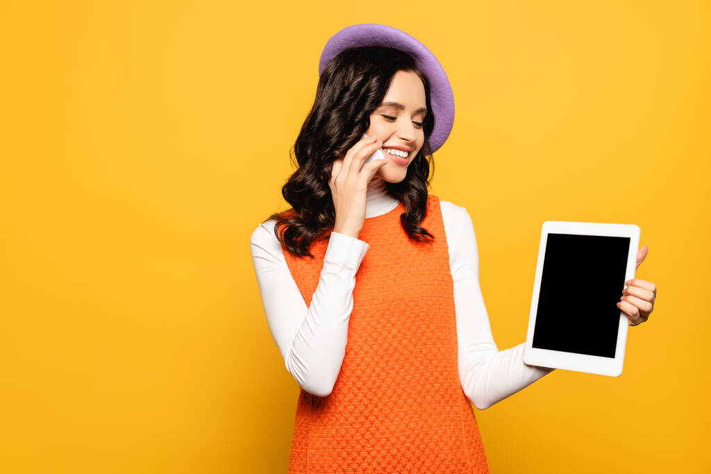 笑顔ブルネット女性でベレー帽話上の携帯電話中に見ますデジタルタブレット絶縁上黄色 - 写真・画像