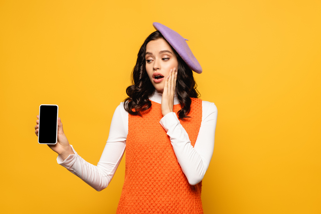 Surprised brunette woman with open mouth showing smartphone with blank screen isolated on yellow - Photo, Image
