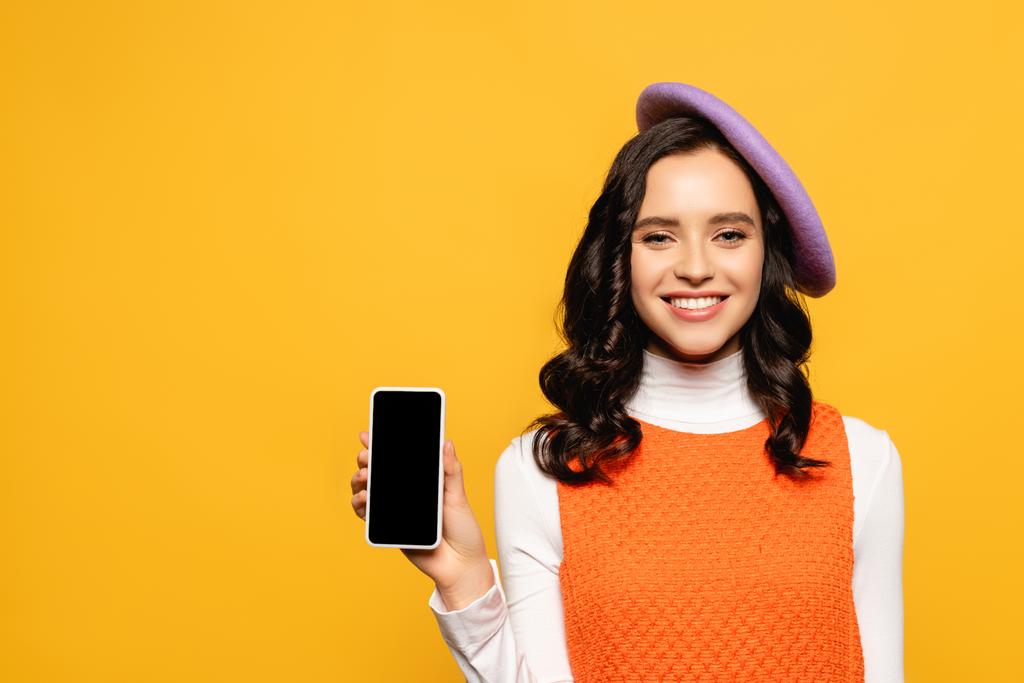 幸せなブルネット女性でベレー帽を見ながらカメラを見せますスマートフォン隔離上の黄色 - 写真・画像