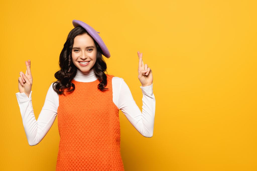 Happy brunette woman in beret with crossed fingers looking at camera isolated on yellow - Photo, Image