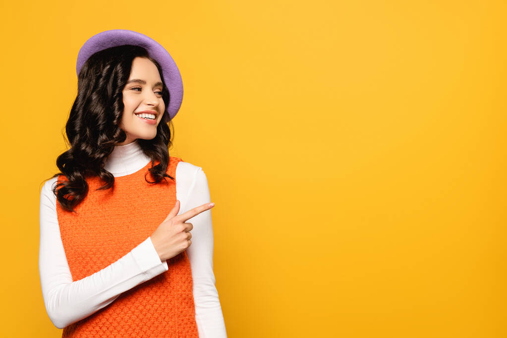 Happy brunette woman in beret looking away while pointing with finger isolated on yellow - Photo, Image