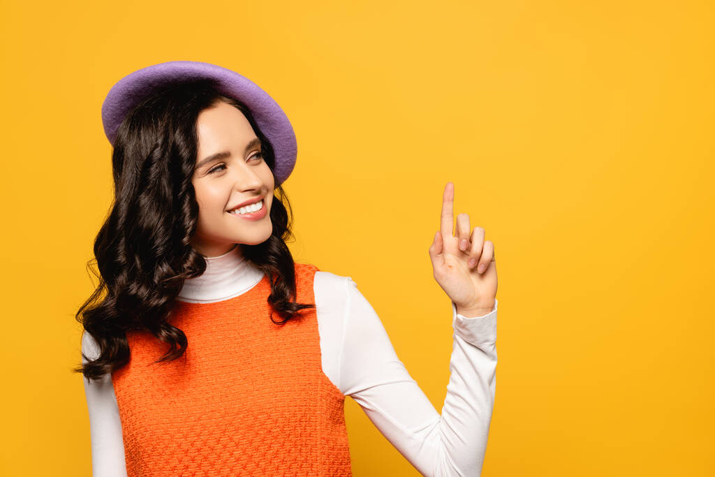 Smiling brunette woman wearing beret and looking away while pointing with finger isolated on yellow - Photo, Image