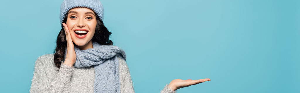 Szczęśliwa brunetka z otwartymi ustami ubrana w strój zimowy i wskazująca ręką odizolowaną na niebiesko, sztandar - Zdjęcie, obraz