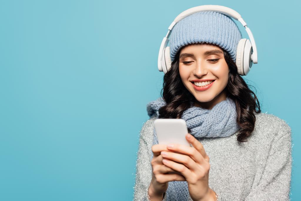 Mulher alegre em roupa de inverno com fones de ouvido mensagens no smartphone isolado em azul - Foto, Imagem