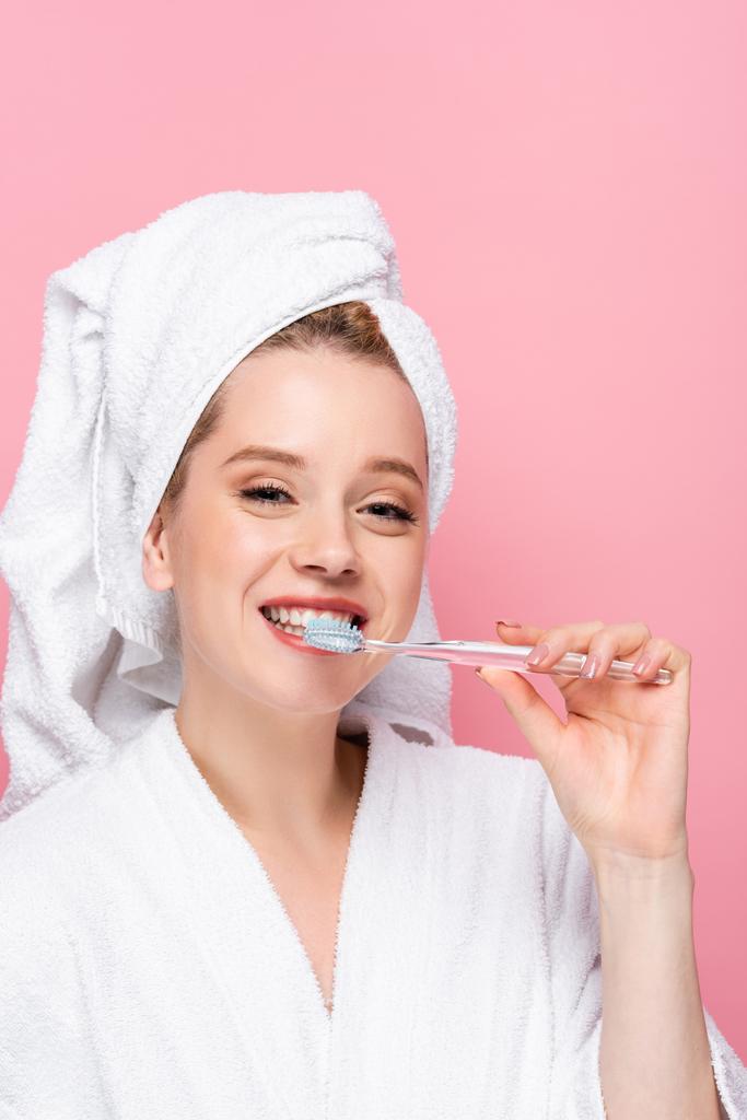 χαμογελαστή νεαρή γυναίκα με μπουρνούζι και πετσέτα στο κεφάλι βουρτσίζοντας τα δόντια που απομονώνονται σε ροζ - Φωτογραφία, εικόνα