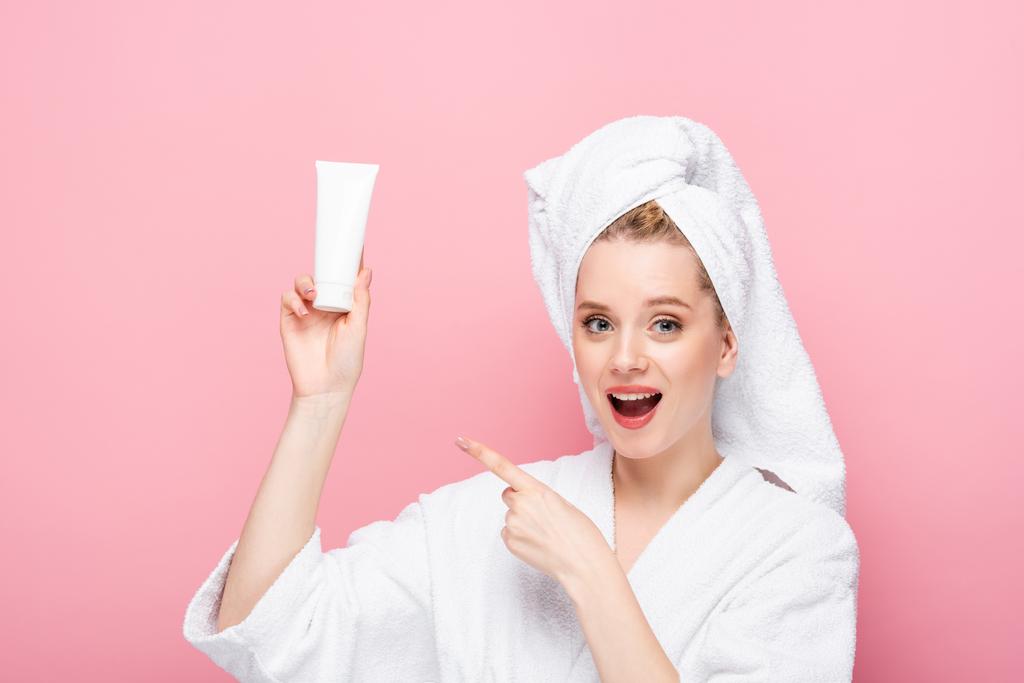 animado jovem mulher em roupão de banho com toalha na cabeça apontando para tubo com creme de mão isolado em rosa - Foto, Imagem