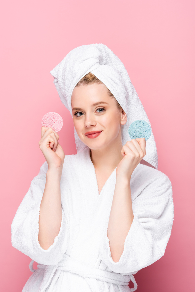 mladá žena v županu s ručníkem na hlavě drží obličejové houby izolované na růžové - Fotografie, Obrázek