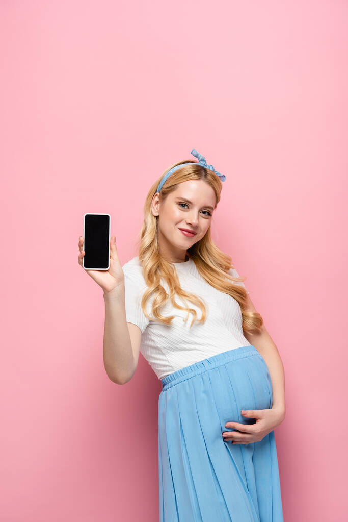 heureuse blonde jeune femme enceinte montrant smartphone sur fond rose - Photo, image