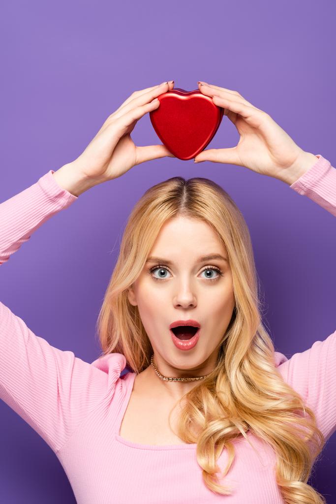 šokované blondýny mladá žena drží červené srdce ve tvaru krabice nad hlavou na fialovém pozadí - Fotografie, Obrázek