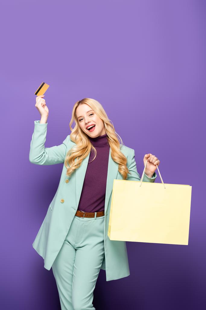 happy blonde young woman in fashionable turquoise blazer with credit card and shopping bag on purple background - Photo, Image