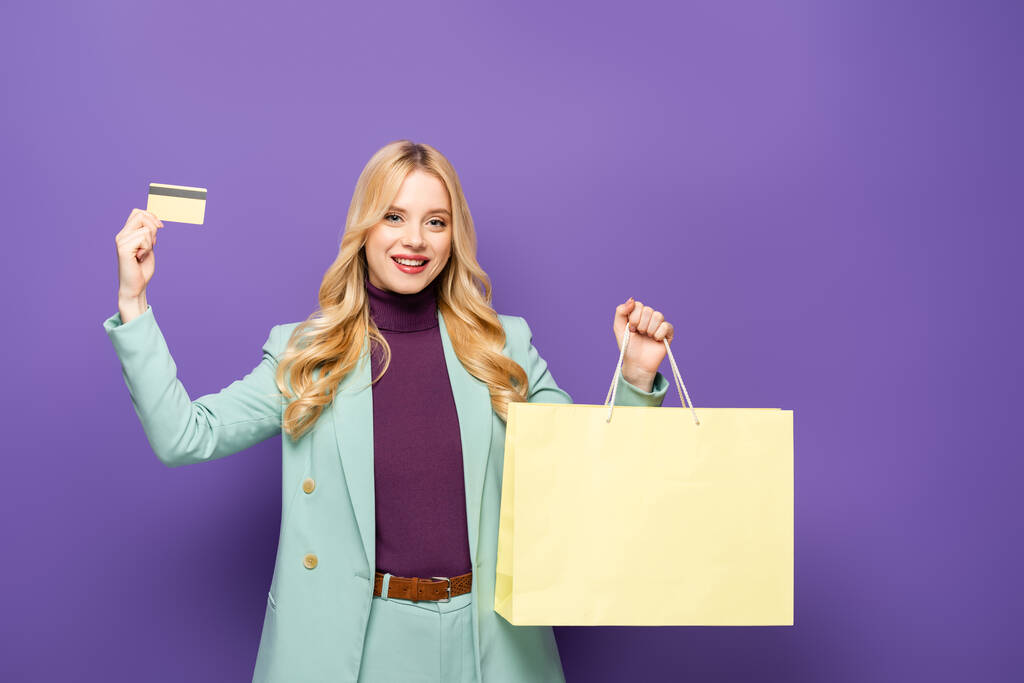 gelukkig blond jong vrouw in modieuze turquoise blazer met credit card en boodschappentas op paarse achtergrond - Foto, afbeelding