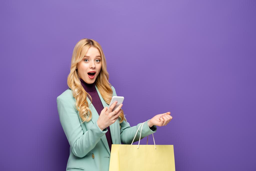 shocked blonde young woman in fashionable turquoise blazer with smartphone and shopping bag on purple background - Photo, Image