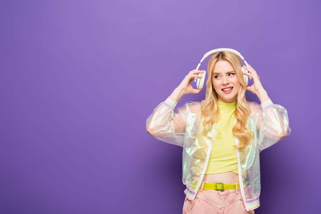 улыбающаяся блондинка в красочном наряде и наушниках на фиолетовом фоне - Фото, изображение