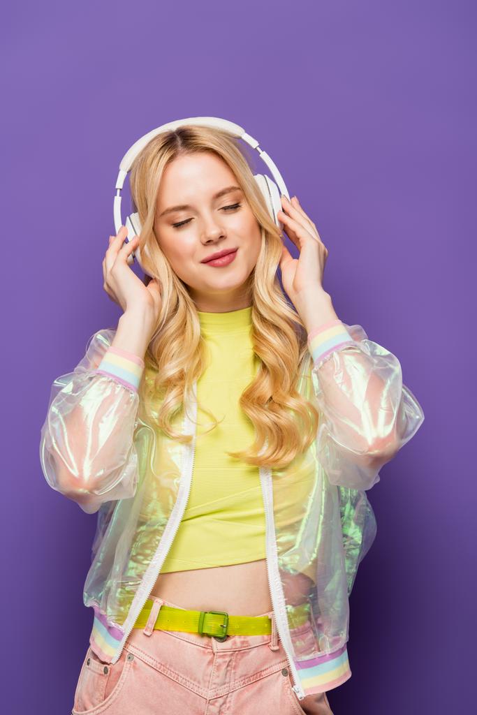 blond jong vrouw in kleurrijke outfit en hoofdtelefoon met gesloten ogen op paarse achtergrond - Foto, afbeelding