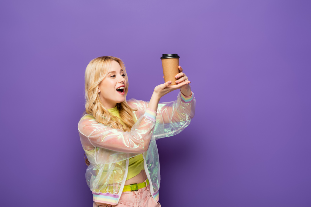 aufgeregte blonde junge Frau in buntem Outfit mit Pappbecher auf lila Hintergrund - Foto, Bild