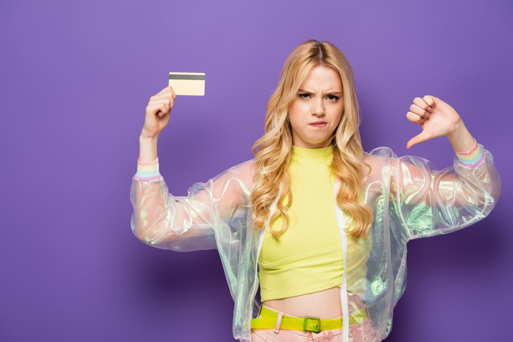 triste jeune femme blonde en tenue colorée montrant carte de crédit et pouce vers le bas sur fond violet - Photo, image
