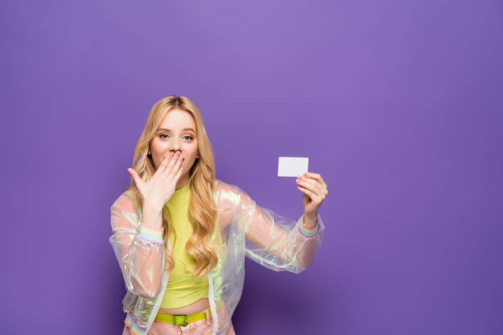järkyttynyt blondi nuori nainen värikäs asu tilalla tyhjä kortti violetti tausta - Valokuva, kuva