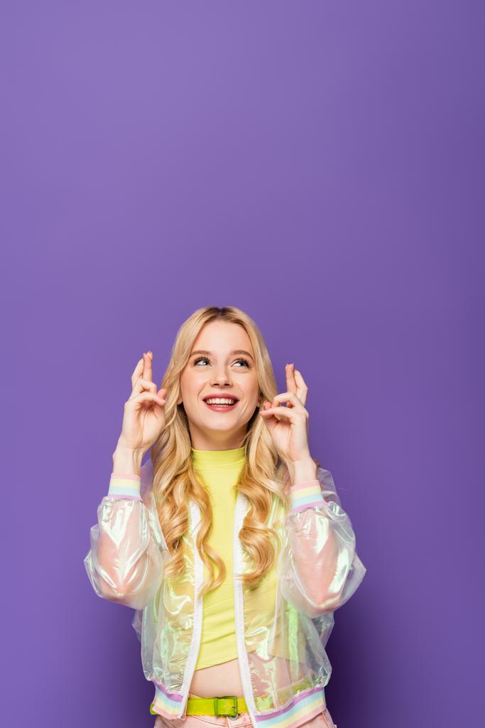 heureuse jeune femme blonde en tenue colorée avec les doigts croisés sur fond violet - Photo, image