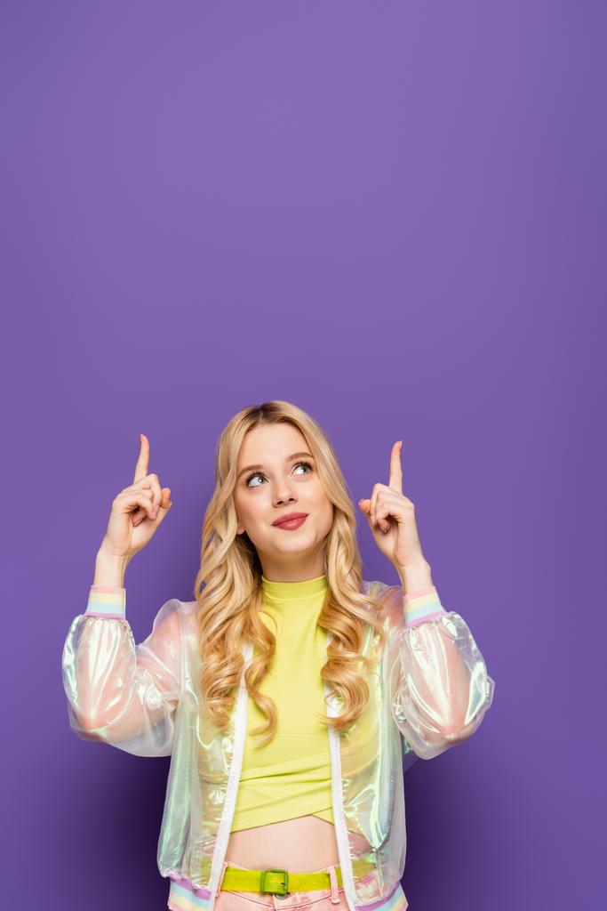 blonde young woman in colorful outfit pointing up on purple background - Photo, Image