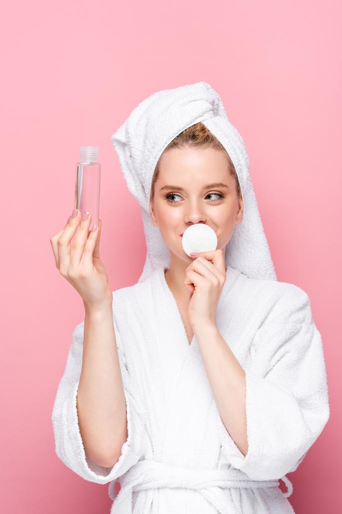 young woman in bathrobe with towel on head with micellar water and cotton pad isolated on pink - Photo, Image