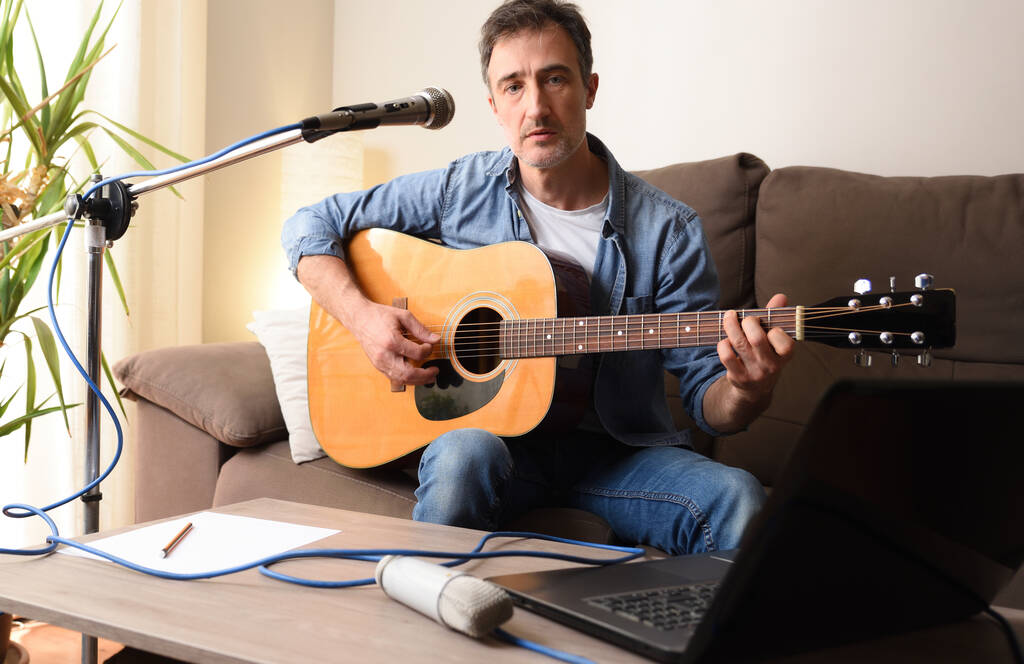 Uomo che suona la chitarra acustica a casa sul divano registrando con apparecchiature per musica con microfono e laptop su tavolo in legno. - Foto, immagini