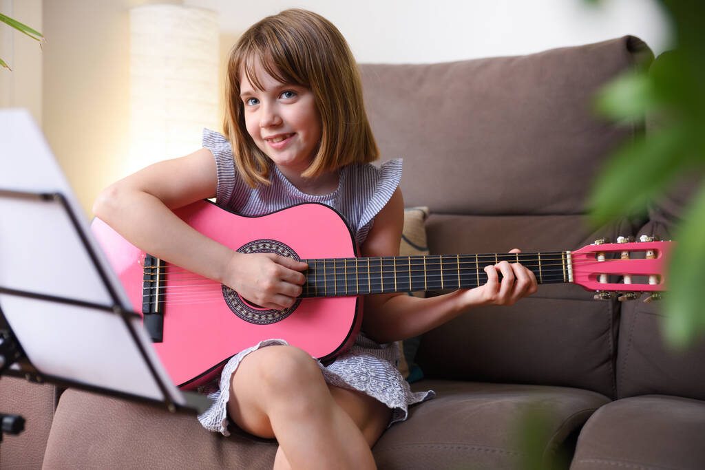笑顔の女の子練習ザギター座っています上のソファで彼女のリビングルーム横に見える - 写真・画像