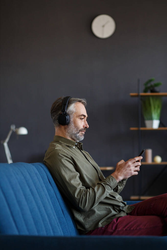 Starszy mężczyzna w słuchawkach słuchający muzyki na smartfonie za pomocą aplikacji muzycznej. Portret przystojnego siwego mężczyzny w słuchawkach i komórce w domu. Relaks, wypoczynek. Miłego słuchania muzyki. - Zdjęcie, obraz