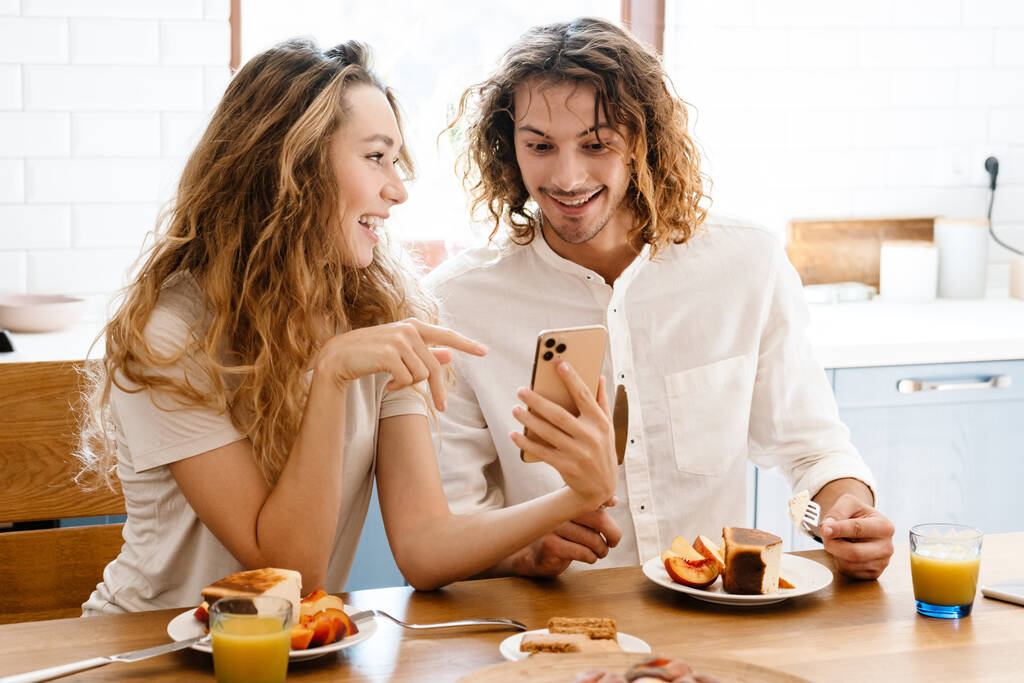 Счастливая красивая пара улыбается и пользуется мобильным телефоном во время завтрака на уютной кухне - Фото, изображение