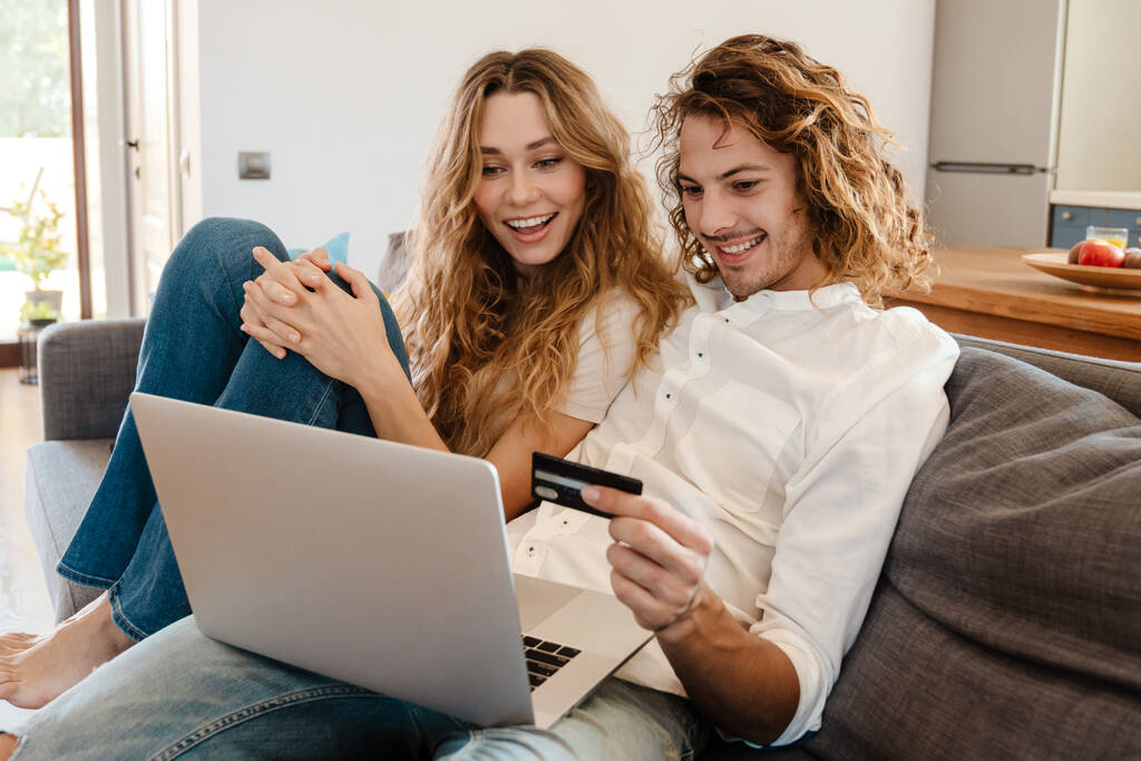 Радостная красивая пара, пользующаяся кредитной картой и ноутбуком, отдыхая дома на диване - Фото, изображение