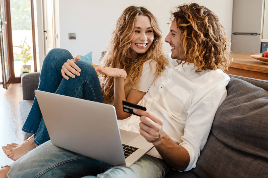 Joyful beautiful couple using credit card and laptop while resting on couch at home - Photo, Image