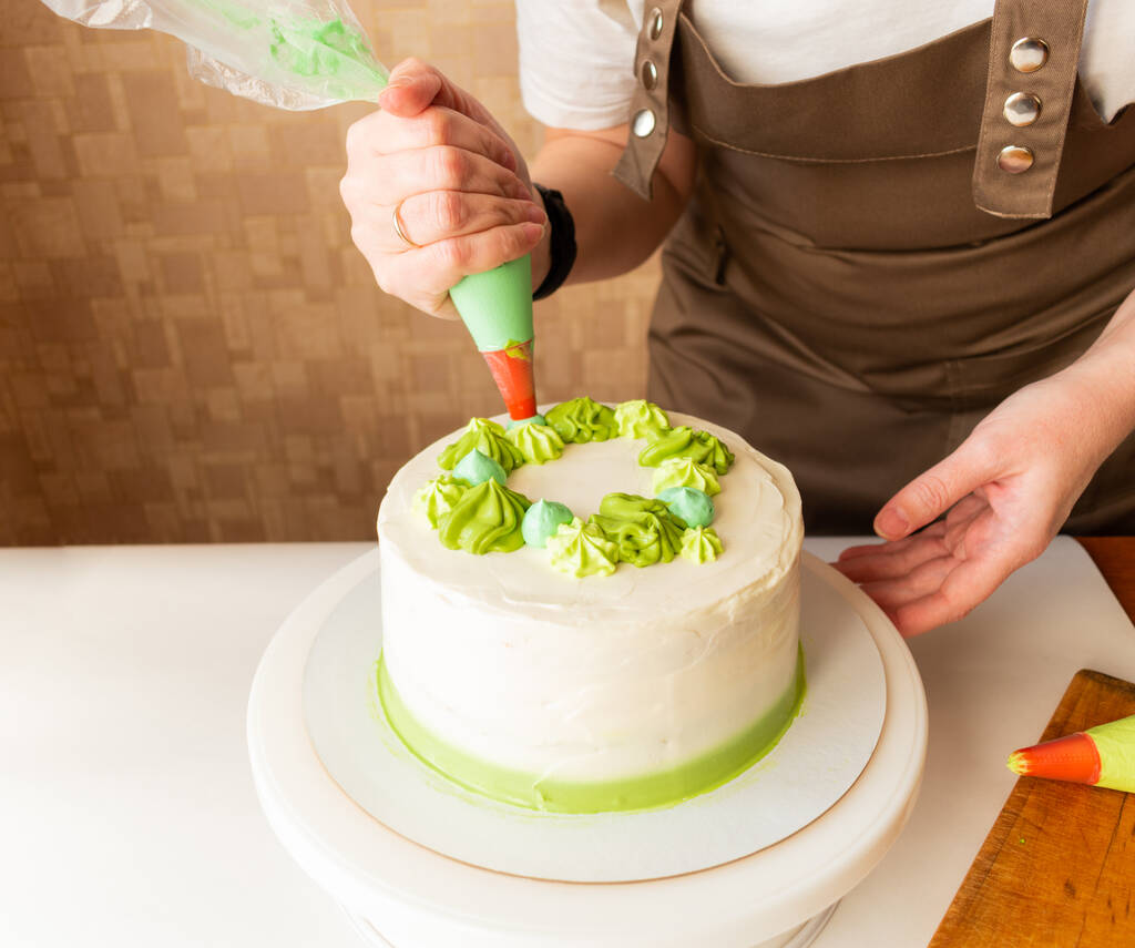 Женщина использует кондитерскую сумку для нанесения взбитых сливок зеленого цвета и украшения именинного торта дома. Концепция домашнего приготовления и украшения торта. Кондитер-женщина делает торт. - Фото, изображение
