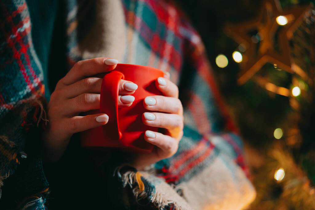 クリスマスツリーの背景に居心地の良い赤いマグカップを持っている女性。熱い飲み物の女の子-お茶、コーヒーまたはココア。新年のコンセプト、快適さと暖かさ. - 写真・画像