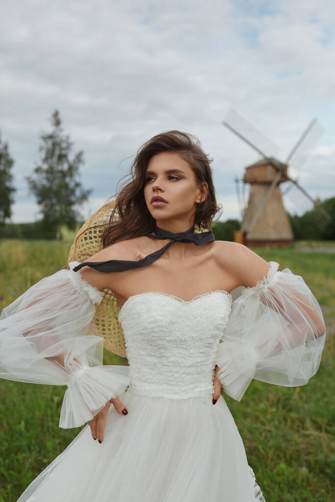 Gyönyörű fiatal menyasszony elegáns ruhában és szalmakalapban pózol a vidéken. Modell lány a vintage esküvői ruha hosszú ujjú - Fotó, kép